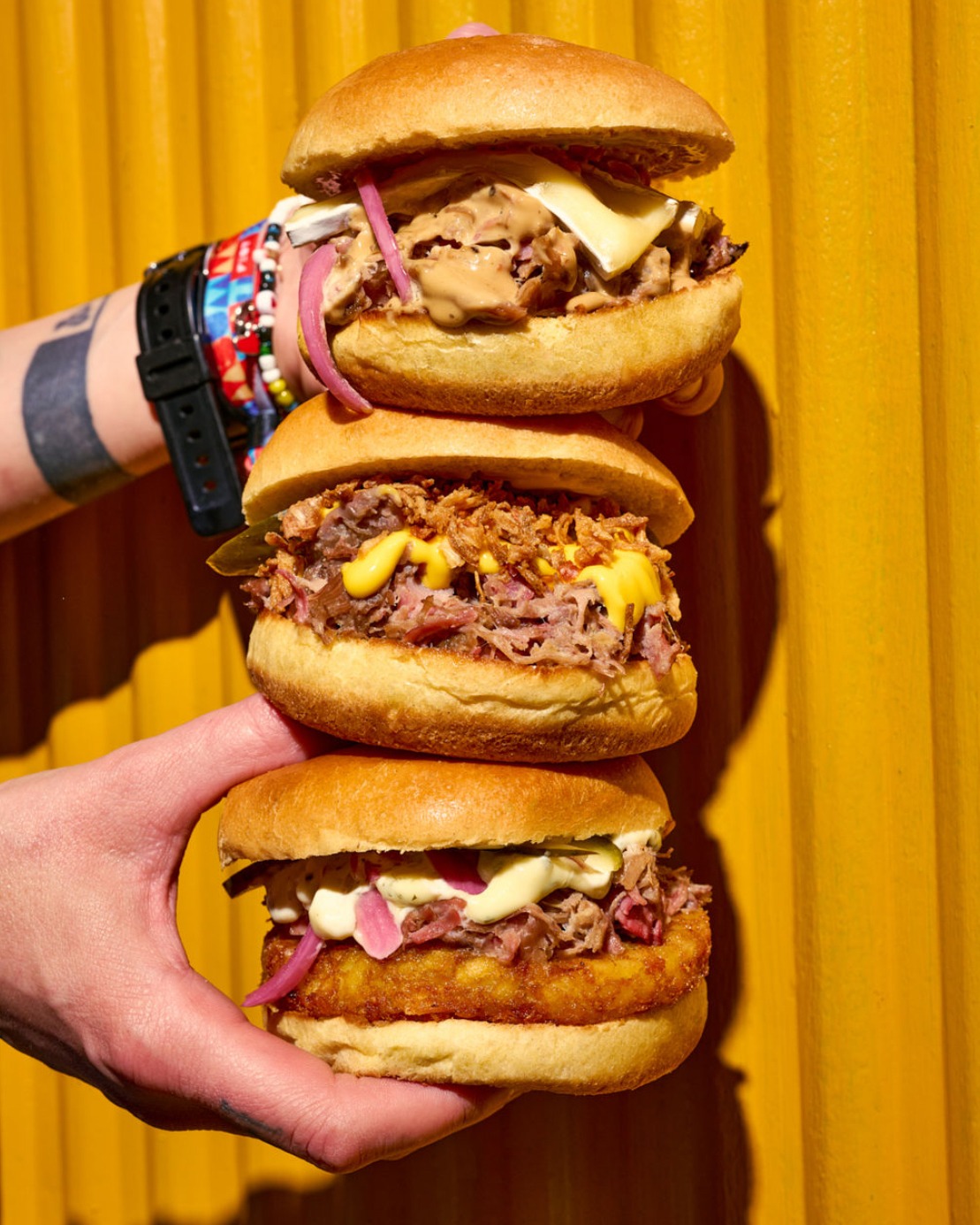 Trois burgers au canard tenus à la main devant un mur jaune